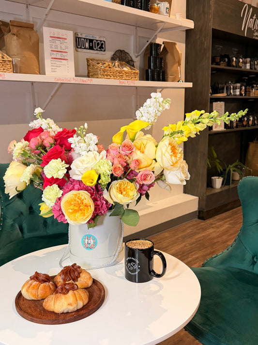 Extra-large luxury floral arrangement in a white signature hat box with yellow garden roses, yellow snapdragons, and a magenta hydrangea on a white cafe table with coffee by Lapin Flowers. Premium elite floral delivery for Potomac MD and Washington DC.⁠