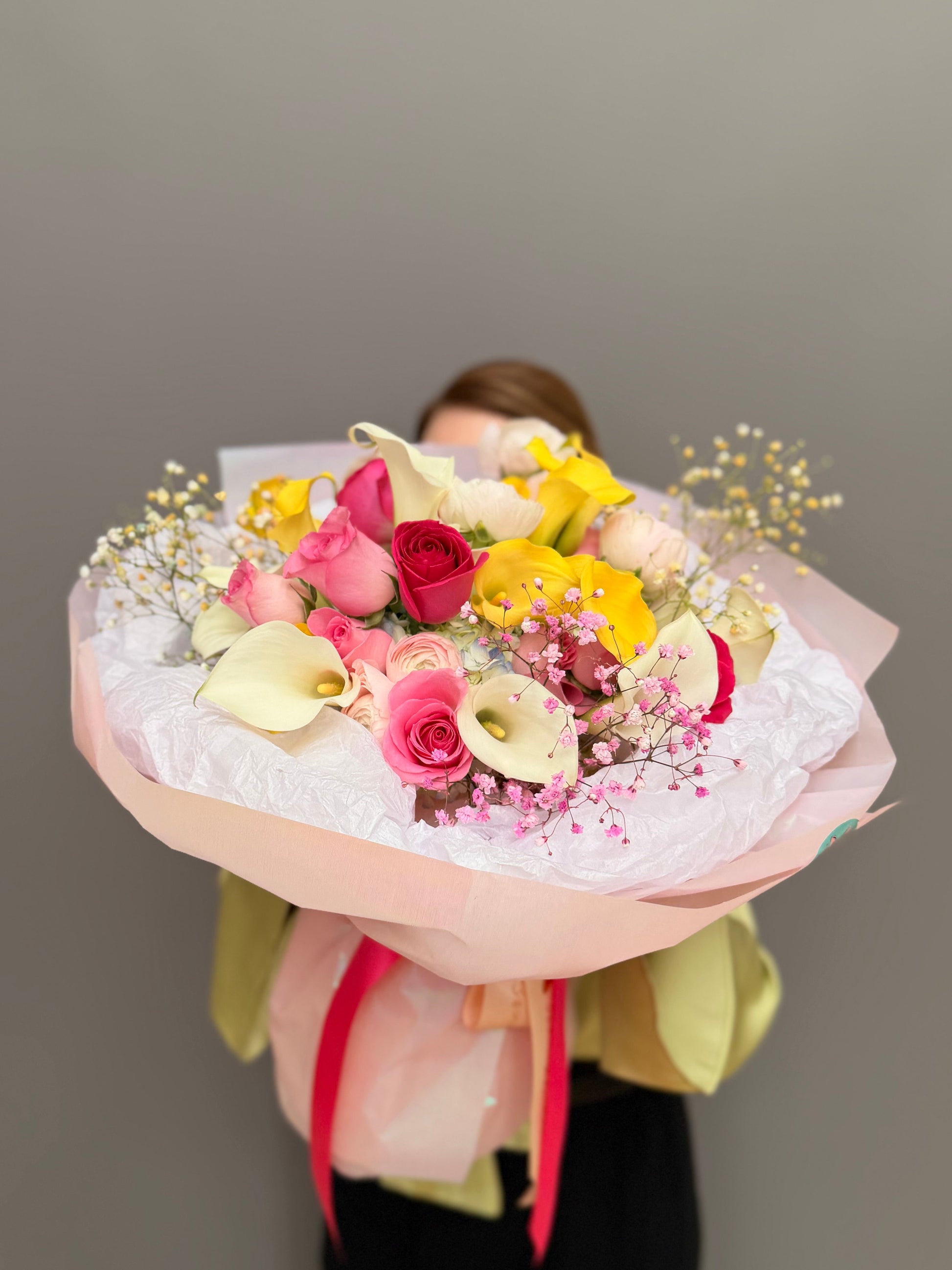 Large luxury mixed bouquet with yellow calla lilies, white calla lilies, magenta roses, and ranunculus wrapped in a big white tissue cloud held by a person by Lapin Flowers. Premium elite event floral design for Washington DC and Potomac MD, McLean VA, Bethesda MD, Annapolis MD. 