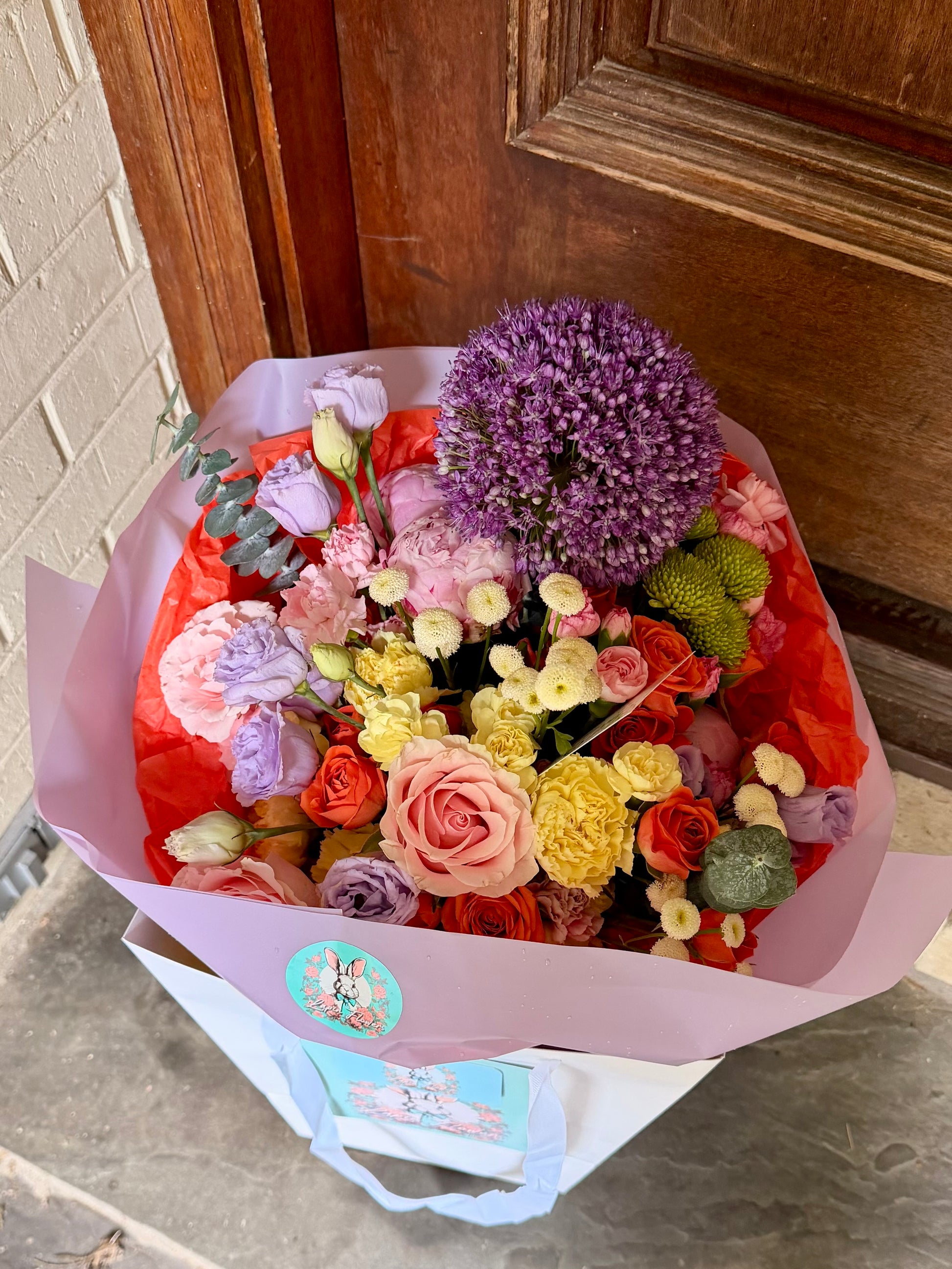 Extra-large luxury artisan bouquet featuring a giant purple Allium, coral and orange roses, lavender lisianthus, and white button flowers wrapped in a vibrant orange and lilac paper by Lapin Flowers. Premium flower delivery in Maryland, Washington DC, and Virginia.⁠
