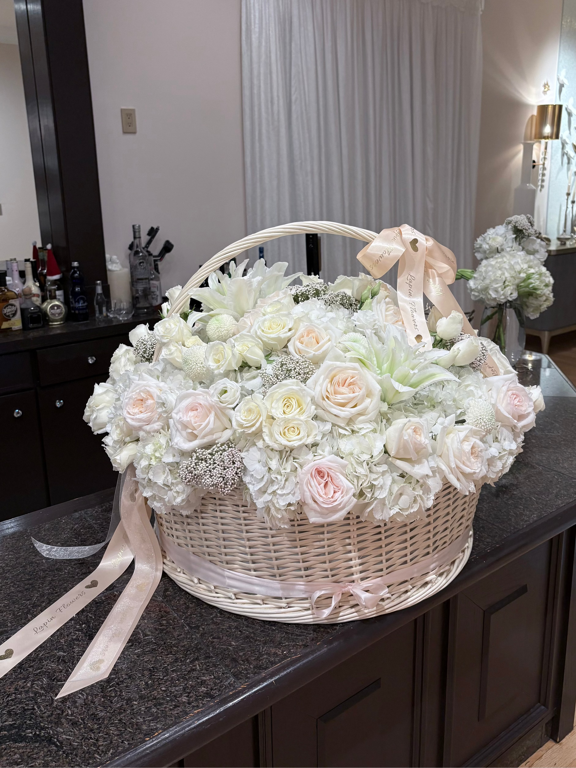 Floral arrangement in a wicker basket on a kitchen counter. Large-scale luxury white flower basket by Lapin Flowers for an engagement in Maryland. Featuring 25 premium Jumbo white hydrangeas, blush O'Hara garden roses, and white lilies in a 24-inch wicker basket. Professional floral installation for upscale events in the MD, VA, and DC area. 