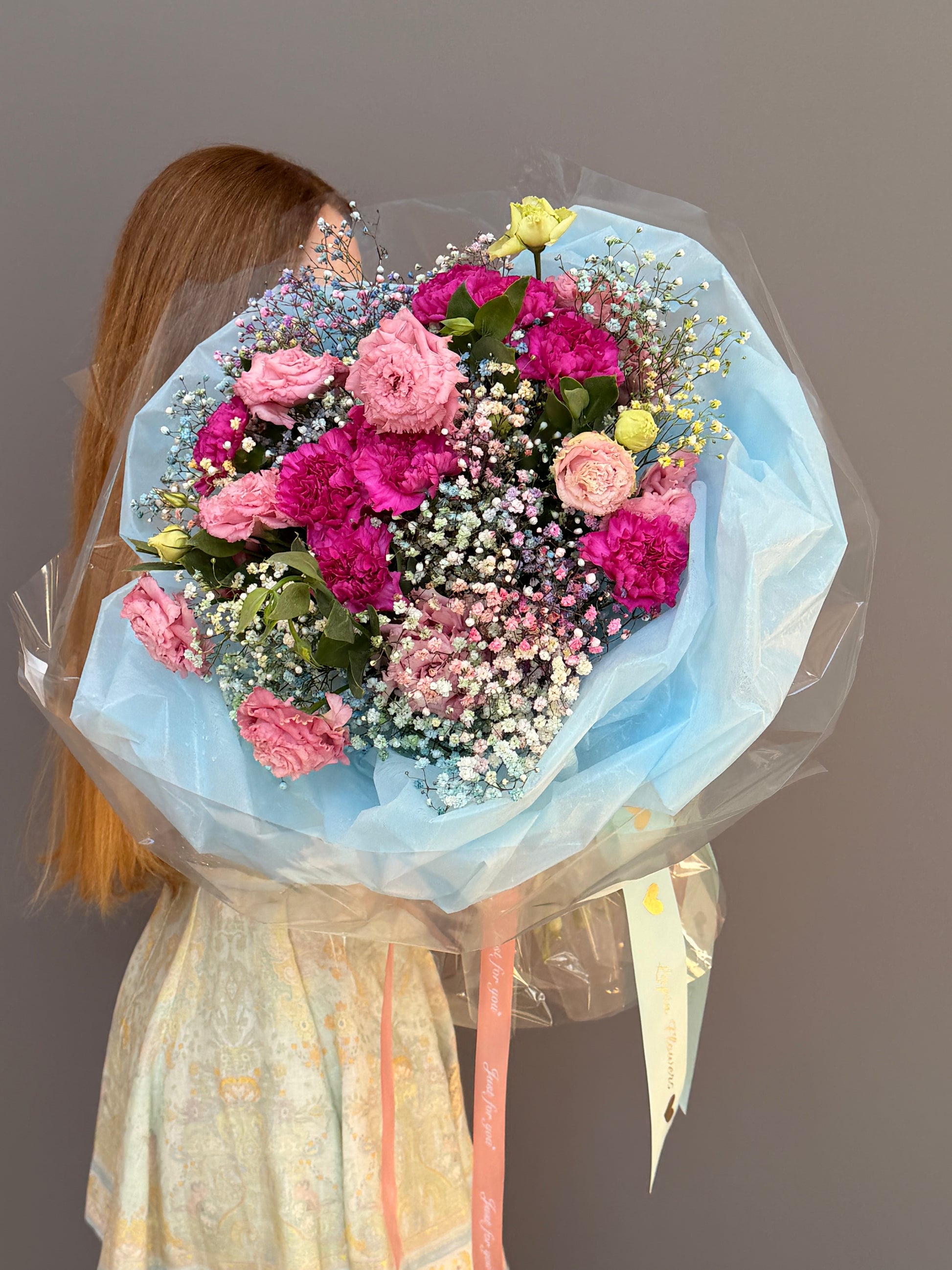 Person holding a Extra-large luxury artisan flower bouquet with tinted blue and pink gypsophila, magenta carnations, and pink lisianthus wrapped in ice-blue paper by Lapin Flowers. Upscale grand delivery for Washington DC, Maryland, and Virginia.