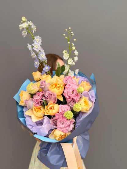A lush, hand-tied bouquet of fresh flowers featuring peach garden roses, lavender roses, pink lisianthus, and tall light blue delphinium spikes. Wrapped in premium blue and lilac paper with a satin ribbon, this luxury floral arrangement is available for delivery in Washington D.C., Arlington, and throughout the DMV area.