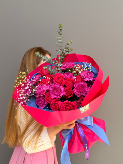 Person holding a bouquet of bright flowers  such as roses and carnationwrapped in hot pink paper with a plain background