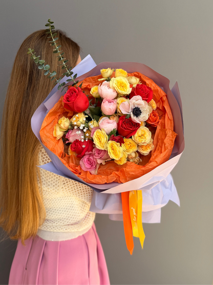 Person holding a colorful bouquet of flowers: Ranunculus, Roses, Anemonies, gypsophila,  with an orange wrap against a plain background