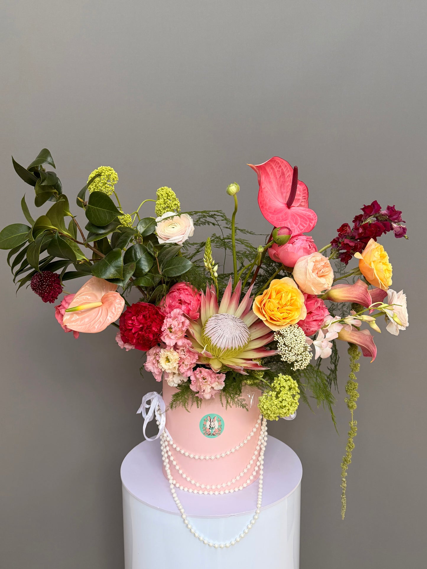 Luxury couture flower arrangement in a pink hatbox featuring a King Protea, coral and pink anthuriums, garden roses, and calla lilies, decorated with elegant pearls by Lapin Flowers, upscale delivery in Potomac Maryland and Washington DC.