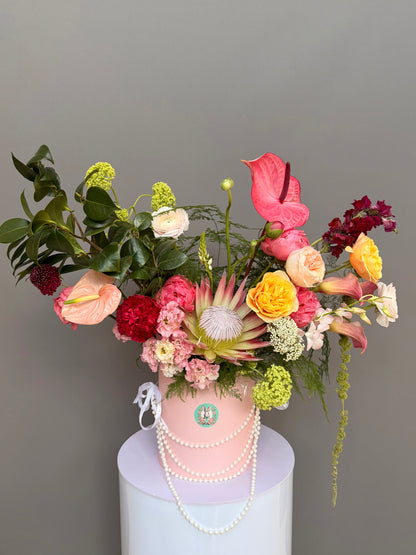 Luxury couture flower arrangement in a pink hatbox featuring a King Protea, coral and pink anthuriums, garden roses, and calla lilies, decorated with elegant pearls by Lapin Flowers, upscale delivery in Potomac Maryland and Washington DC.