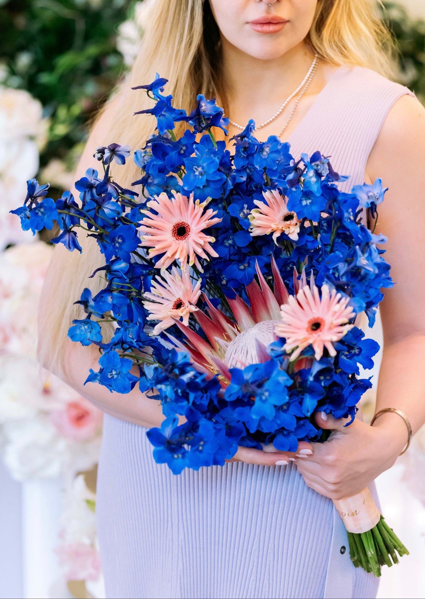 Luxury contemporary artisan bouquet with a large pink King Protea, blush spider gerberas, and a dramatic cloud of electric blue delphinium held by a florist by Lapin Flowers. Premium exotic flower delivery for Washington DC and Maryland.⁠