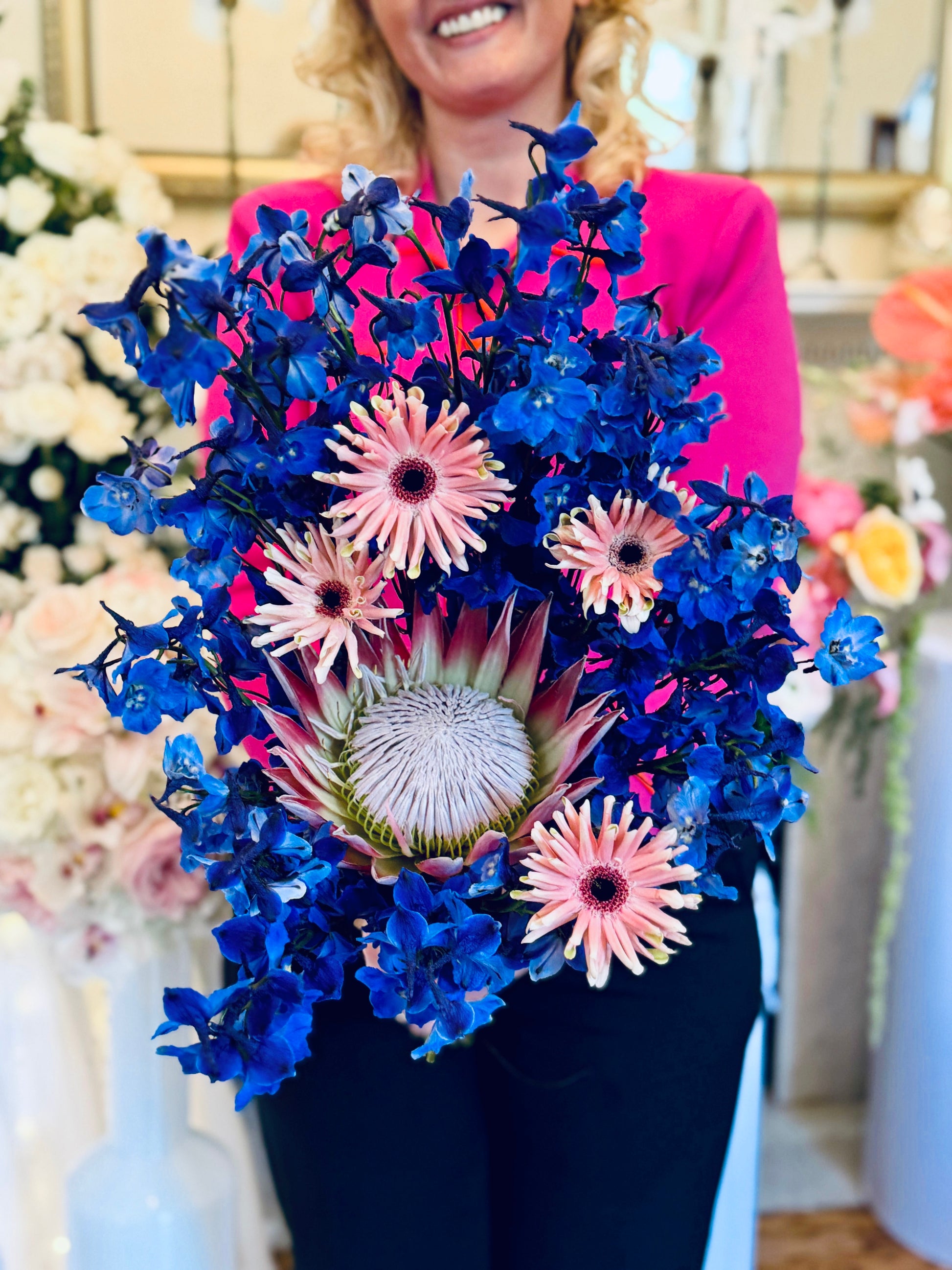Luxury contemporary artisan bouquet with a large pink King Protea, blush spider gerberas, and a dramatic cloud of electric blue delphinium held by a florist by Lapin Flowers. Premium exotic flower delivery for Washington DC and Maryland.⁠