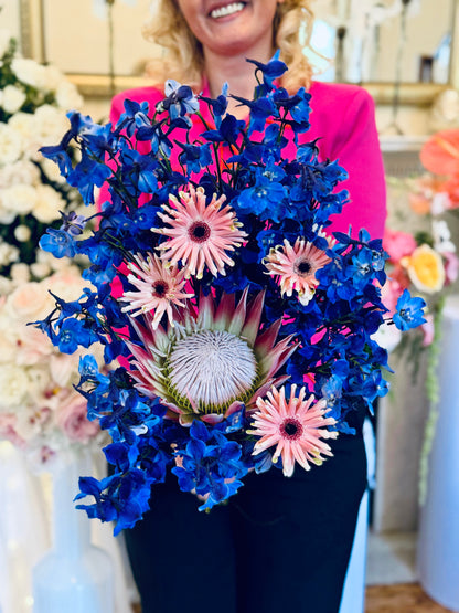 Luxury contemporary artisan bouquet with a large pink King Protea, blush spider gerberas, and a dramatic cloud of electric blue delphinium held by a florist by Lapin Flowers. Premium exotic flower delivery for Washington DC and Maryland.⁠