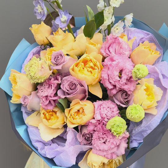 A lush, hand-tied bouquet of fresh flowers featuring peach garden roses, lavender roses, pink lisianthus, and tall light blue delphinium spikes. Wrapped in premium blue and lilac paper with a satin ribbon, this luxury floral arrangement is available for delivery in Washington D.C., Arlington, and throughout the DMV area.