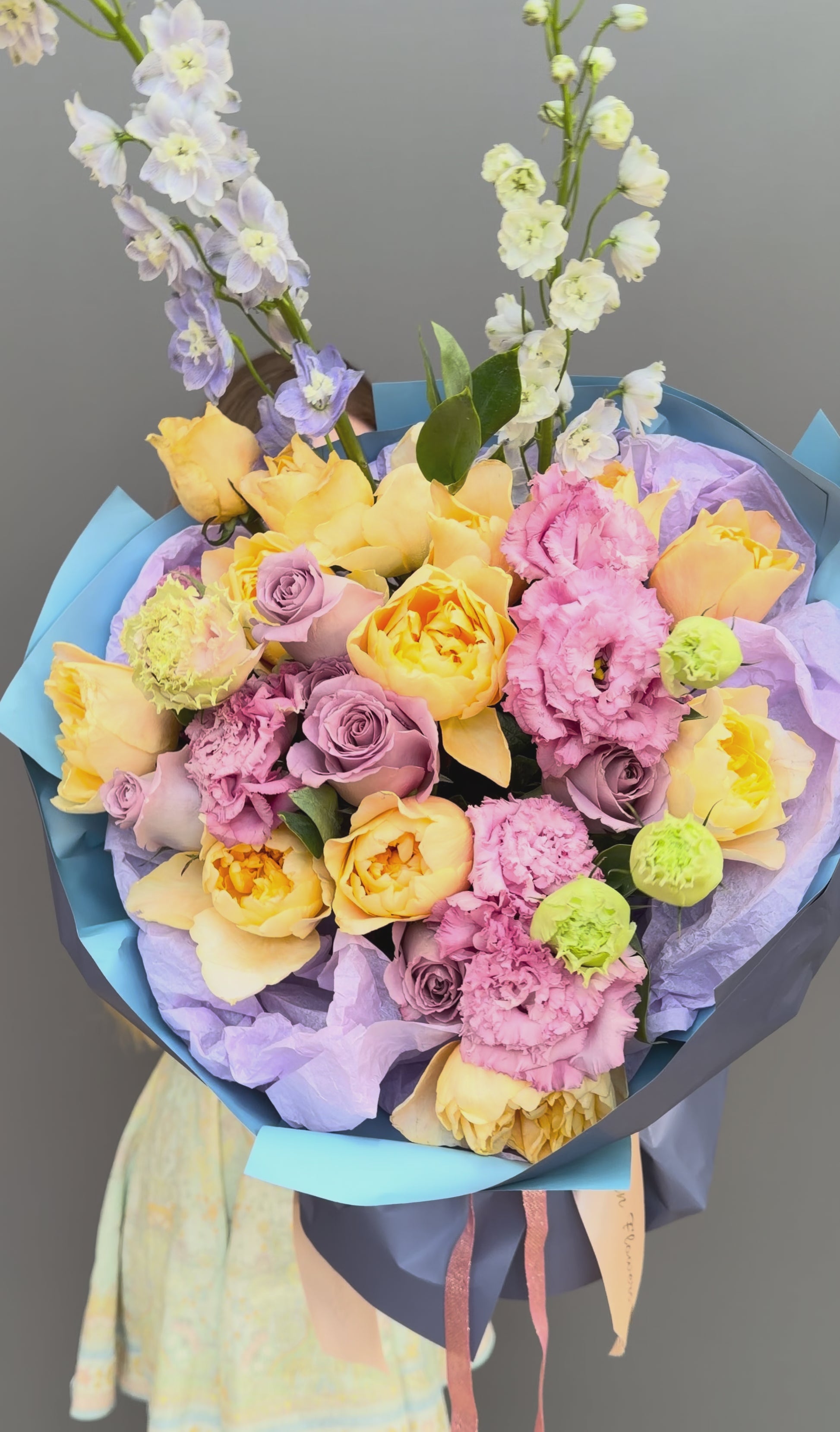 A lush, hand-tied bouquet of fresh flowers featuring peach garden roses, lavender roses, pink lisianthus, and tall light blue delphinium spikes. Wrapped in premium blue and lilac paper with a satin ribbon, this luxury floral arrangement is available for delivery in Washington D.C., Arlington, and throughout the DMV area.