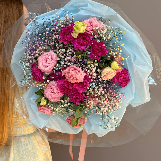 Person holding a Extra-large luxury artisan flower bouquet with tinted blue and pink gypsophila, magenta carnations, and pink lisianthus wrapped in ice-blue paper by Lapin Flowers. Upscale grand delivery for Washington DC, Maryland, and Virginia.