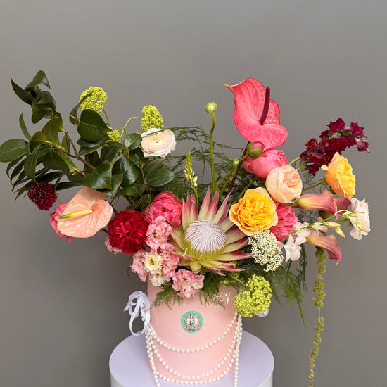 Luxury couture flower arrangement in a pink hatbox featuring a King Protea, coral and pink anthuriums, garden roses, and calla lilies, decorated with elegant pearls by Lapin Flowers, upscale delivery in Potomac Maryland and Washington DC.